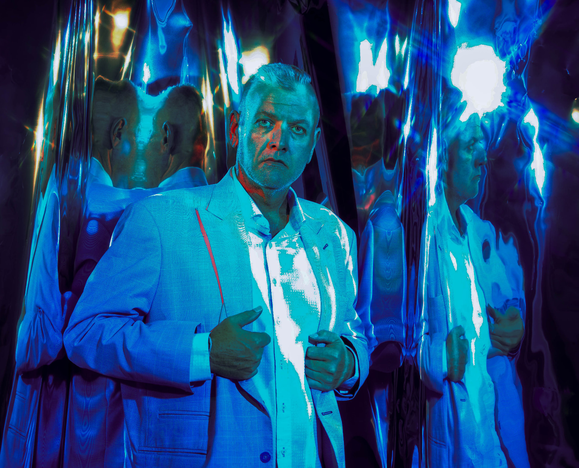 A figure in a blue suit standing among angled mirrors, lit from one side so half the frame falls into shadow