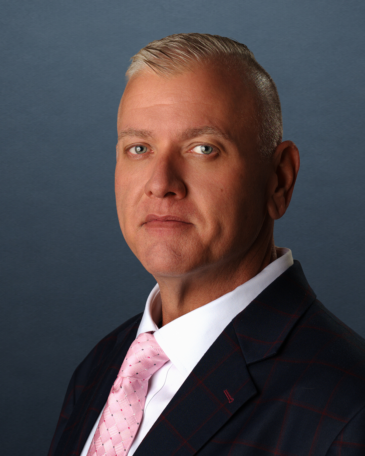 Directed professional headshot of a man in a plaid suit and pink tie against a blue-gray ground