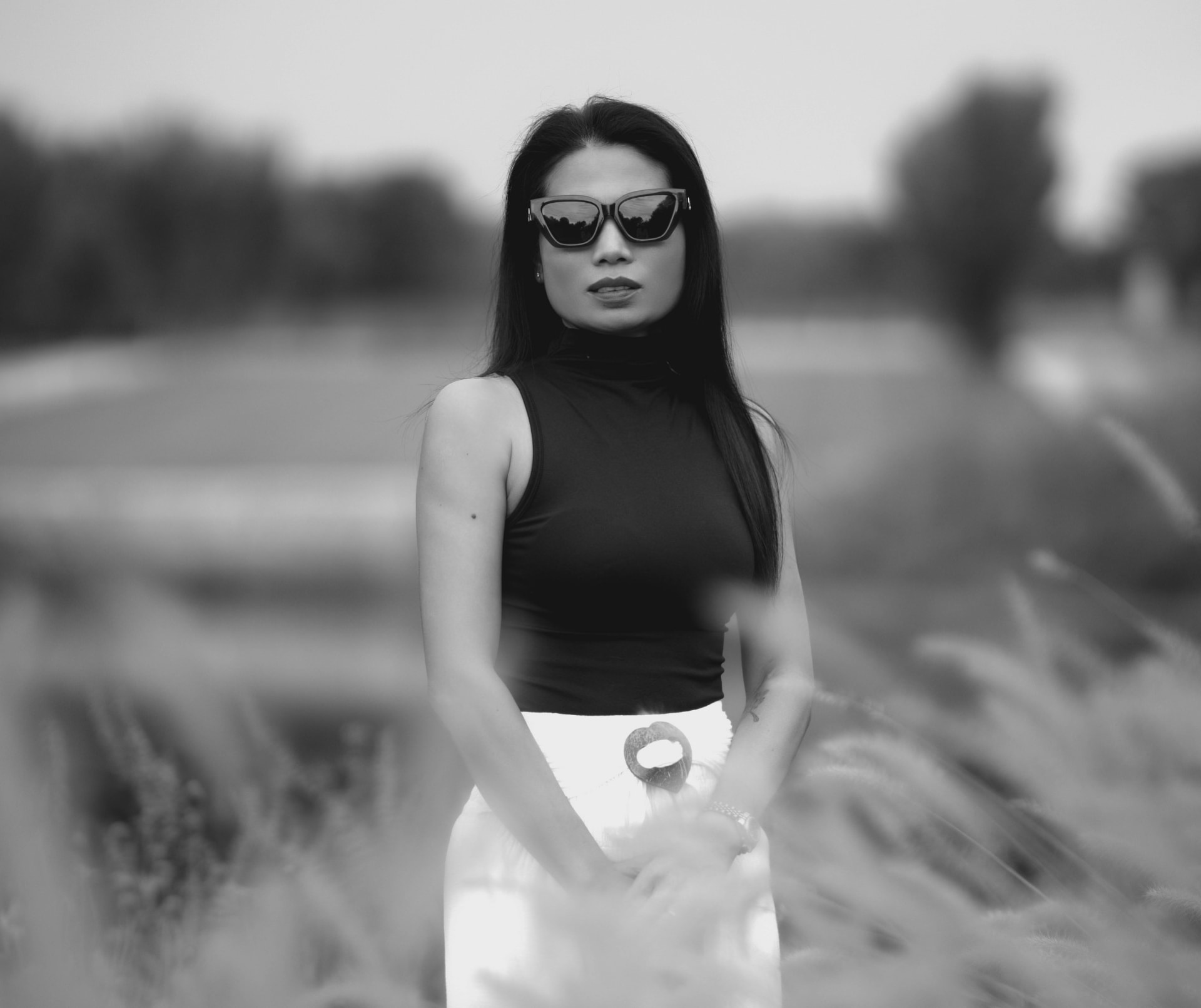 Black-and-white portrait in dark sunglasses and a black sleeveless top, standing in a field of tall grasses
